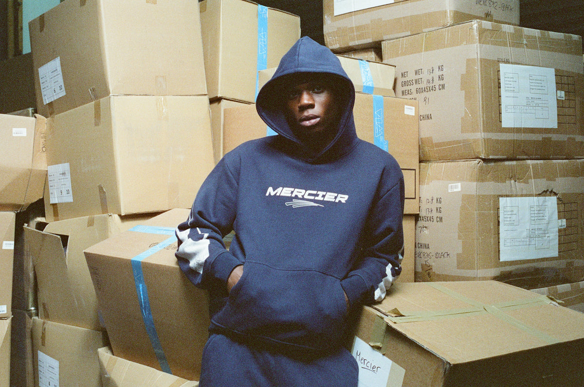 Navy Mercier Speed hoodie worn hood up, in the warehouse among stacked boxes